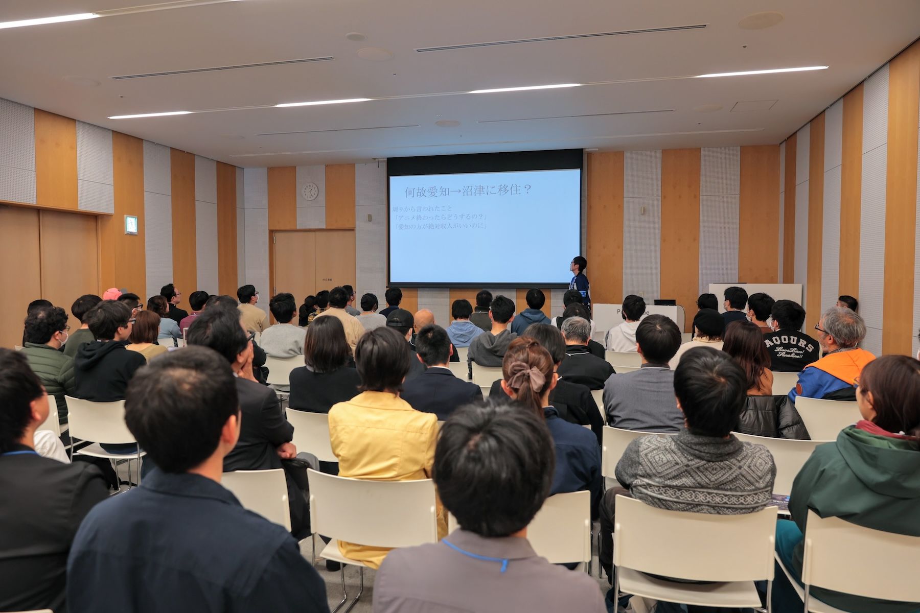 プレゼン資料が映し出されたスクリーンを含む会場全体が写っていおり、座席は満席に近い状態になっている。その中で1人の男性がプレゼン発表している。